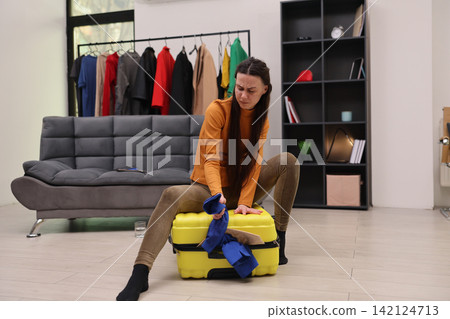 Frustrated woman sitting on yellow suitcase while unpacking clothes in modern living room, surrounded by hanging garments, sofa, shelving, and home interior details, suggesting moving, relocation Frustrated woman sitting on yellow suitcase while unpacking clothes in modern living room, surrounded by hanging garments, sofa, shelving, and home interior details, suggesting moving, relocation 142124713