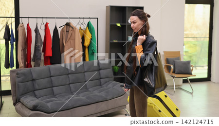Young woman with suitcase standing inside modern fashion showroom beside clothing rack, gray sofa, display shelving, and bright natural window light, browsing stylish outfits Young woman with suitcase standing inside modern fashion showroom beside clothing rack, gray sofa, display shelving, and bright natural window light, browsing stylish outfits 142124715