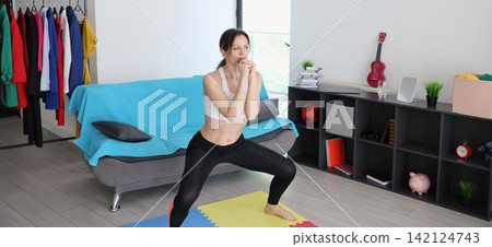 Woman practicing yoga squat pose in bright living room with fitness mat, casual home workout setup, colorful clothes rack, sofa, shelving, and thoughtful focused expression during indoor exercise 142124743
