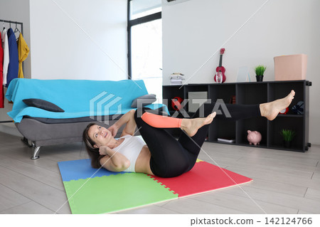 Woman doing bicycle crunch exercise on colorful mat in bright home living room with sofa, shelves, clothes rack, fitness accessories, and natural daylight creating healthy active lifestyle scene Woman doing bicycle crunch exercise on colorful mat in bright home living room with sofa, shelves, clothes rack, fitness accessories, and natural daylight creating healthy active lifestyle scene 142124766