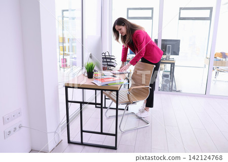 Businesswoman arranging papers at desk in bright modern office while standing beside computer chair near glass walls, organized workspace, focused planning, administrative work, productivity Businesswoman arranging papers at desk in bright modern office while standing beside computer chair near glass walls, organized workspace, focused planning, administrative work, productivity 142124768
