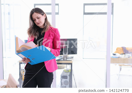 Businesswoman in pink blazer reviewing documents in bright modern office while holding blue folder and standing near computer workstation, focused on paperwork and office tasks Businesswoman in pink blazer reviewing documents in bright modern office while holding blue folder and standing near computer workstation, focused on paperwork and office tasks 142124770