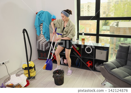 Young woman cleaning modern apartment with mop and dustpan beside storage shelf, vacuum, laundry basket, and large city window, showing domestic chores and everyday home maintenance Young woman cleaning modern apartment with mop and dustpan beside storage shelf, vacuum, laundry basket, and large city window, showing domestic chores and everyday home maintenance 142124876