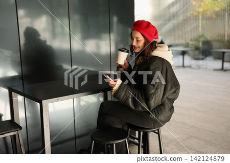 Young woman in red beret sits at modern cafe table, smiling at smartphone while holding takeaway coffee cup, wearing dark coat and scarf beside reflective wall in soft daylight Young woman in red beret sits at modern cafe table, smiling at smartphone while holding takeaway coffee cup, wearing dark coat and scarf beside reflective wall in soft daylight 142124879
