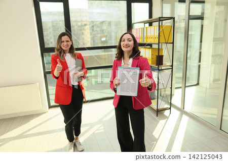 Two smiling businesswomen standing in bright modern office, holding clipboard documents and giving thumbs up beside large windows, reflecting teamwork, approval, success, confident professional 142125043