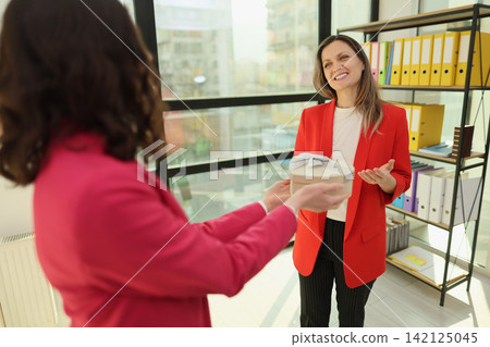 Smiling businesswoman in red blazer receives gift box from colleague during office celebration beside bright window and shelves with colorful binders in modern workplace Smiling businesswoman in red blazer receives gift box from colleague during office celebration beside bright window and shelves with colorful binders in modern workplace 142125045