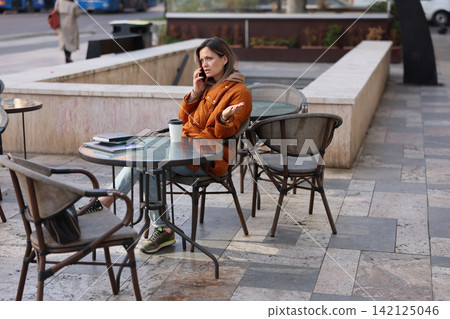 Young woman in orange quilted jacket talking on phone while sitting at outdoor cafe table with coffee notebook and laptop, urban sidewalk setting, thoughtful expression, casual city break 142125046