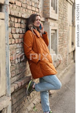 Young woman in orange quilted coat leans against weathered brick wall, talking on smartphone beside narrow urban street with relaxed posture, casual jeans, and soft daylight Young woman in orange quilted coat leans against weathered brick wall, talking on smartphone beside narrow urban street with relaxed posture, casual jeans, and soft daylight 142125051