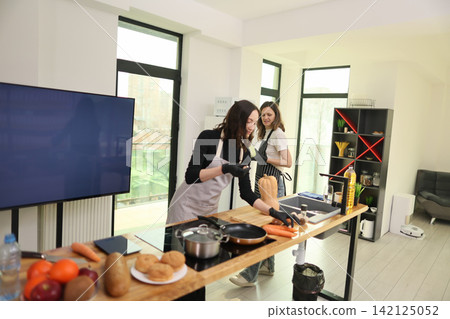 Two women preparing fresh vegetables in bright modern kitchen with island countertop, cookware, sink, open shelving, natural daylight, home cooking, meal preparation, healthy food, teamwork Two women preparing fresh vegetables in bright modern kitchen with island countertop, cookware, sink, open shelving, natural daylight, home cooking, meal preparation, healthy food, teamwork 142125052