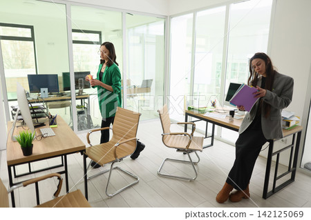Two professional women organizing paperwork in bright modern office with glass walls, desktop computers, office chairs, plants, and sunlight creating focused business workspace Two professional women organizing paperwork in bright modern office with glass walls, desktop computers, office chairs, plants, and sunlight creating focused business workspace 142125069