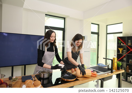 Two women preparing fresh vegetables and cookware on bright modern kitchen island, wearing aprons and gloves while cooking together in stylish home interior with natural daylight Two women preparing fresh vegetables and cookware on bright modern kitchen island, wearing aprons and gloves while cooking together in stylish home interior with natural daylight 142125070