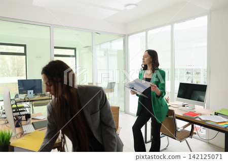 Two businesswomen collaborating in bright modern office, one standing with reports beside desk while colleague works nearby, glass walls, computers, daylight, professional atmosphere Two businesswomen collaborating in bright modern office, one standing with reports beside desk while colleague works nearby, glass walls, computers, daylight, professional atmosphere 142125071