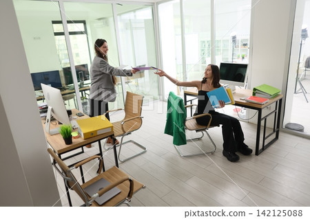 Two women exchanging office files in bright modern workspace, seated colleague holding colorful folders while standing coworker hands over document, glass wall meeting room and desktop computers Two women exchanging office files in bright modern workspace, seated colleague holding colorful folders while standing coworker hands over document, glass wall meeting room and desktop computers 142125088