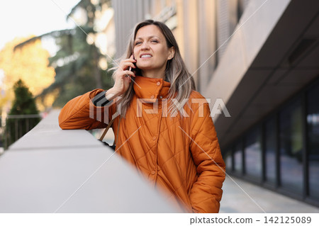 Woman in orange quilted coat talking on smartphone beside modern building outdoors, with autumn light, soft background blur, confident expression, urban lifestyle setting, casual city communication Woman in orange quilted coat talking on smartphone beside modern building outdoors, with autumn light, soft background blur, confident expression, urban lifestyle setting, casual city communication 142125089