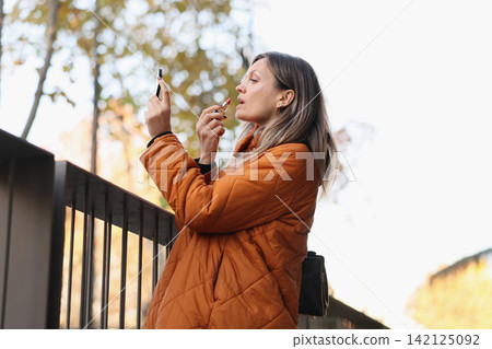 Woman in orange quilted coat applying lipstick while checking compact mirror outdoors beside railing with soft autumn bokeh background and bright natural daylight 142125092