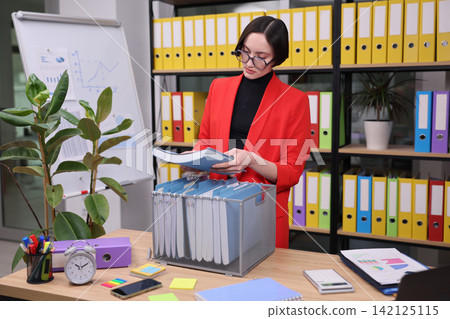 Focused woman in red blazer reviewing files at modern office desk with folders, charts, plants, laptop, and organized shelving, showing business planning, administration, and document management Focused woman in red blazer reviewing files at modern office desk with folders, charts, plants, laptop, and organized shelving, showing business planning, administration, and document management 142125115