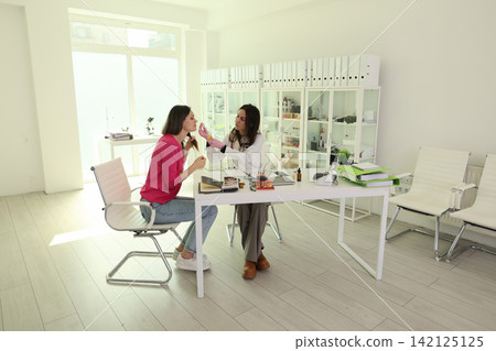 Young woman receiving facial skin examination from specialist in bright medical office, with consultation table, glass cabinets, chairs, and soft daylight creating clean clinical atmosphere Young woman receiving facial skin examination from specialist in bright medical office, with consultation table, glass cabinets, chairs, and soft daylight creating clean clinical atmosphere 142125125