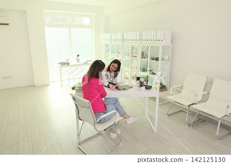 Woman consulting smiling doctor in bright modern office, seated across desk during friendly medical meeting with files, cabinet shelves, chairs, laptop, natural daylight, clean professional interior Woman consulting smiling doctor in bright modern office, seated across desk during friendly medical meeting with files, cabinet shelves, chairs, laptop, natural daylight, clean professional interior 142125130