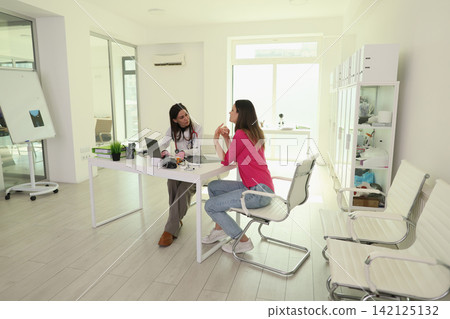Female doctor consulting with young woman in bright modern medical office, seated across desk during face to face conversation, clinical workspace with laptop, chairs, cabinets, daylight Female doctor consulting with young woman in bright modern medical office, seated across desk during face to face conversation, clinical workspace with laptop, chairs, cabinets, daylight 142125132