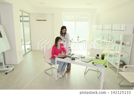 Female doctor showing medical images to seated patient in bright modern consultation office with desk paperwork, glass cabinets, sunlight, and clean minimalist interior for healthcare discussion Female doctor showing medical images to seated patient in bright modern consultation office with desk paperwork, glass cabinets, sunlight, and clean minimalist interior for healthcare discussion 142125133