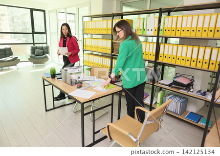 Two professional women reviewing paperwork in bright modern office with shelves of yellow binders, desk laptop, scattered documents, and sunlight creating organized business workspace Two professional women reviewing paperwork in bright modern office with shelves of yellow binders, desk laptop, scattered documents, and sunlight creating organized business workspace 142125134