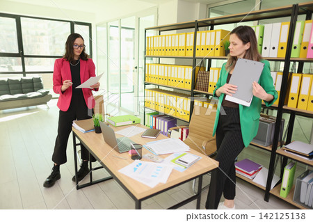 Two businesswomen reviewing documents in bright modern office with organized shelves, laptop, desk papers, and colorful binders, discussing planning, administration, and teamwork during workday Two businesswomen reviewing documents in bright modern office with organized shelves, laptop, desk papers, and colorful binders, discussing planning, administration, and teamwork during workday 142125138