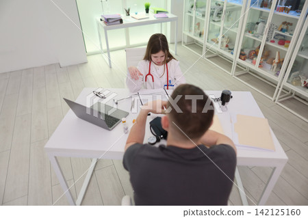 Young girl dressed as doctor examining boy across white desk in bright clinic room with laptop, medical tools, cabinet shelves, and calm professional role play setting Young girl dressed as doctor examining boy across white desk in bright clinic room with laptop, medical tools, cabinet shelves, and calm professional role play setting 142125160