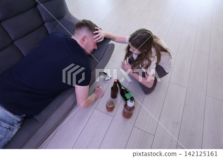 Young girl kneels beside exhausted man on sofa, touching his forehead while empty bottles and spilled drink rest on floor in bright room, suggesting concern, recovery, and household crisis Young girl kneels beside exhausted man on sofa, touching his forehead while empty bottles and spilled drink rest on floor in bright room, suggesting concern, recovery, and household crisis 142125161