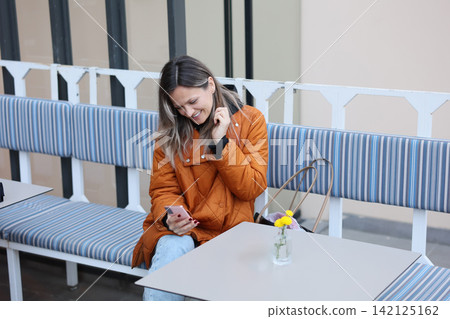 Smiling woman in orange quilted coat using smartphone while seated on striped cafe bench beside small flowers on table in bright outdoor seating area, with woman, smartphone, cafe, sitting detail Smiling woman in orange quilted coat using smartphone while seated on striped cafe bench beside small flowers on table in bright outdoor seating area, with woman, smartphone, cafe, sitting detail 142125162