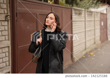 Young woman in black coat holding smartphone near residential wall, standing outdoors on quiet street with thoughtful expression, shoulder bag, soft natural light and shallow focus Young woman in black coat holding smartphone near residential wall, standing outdoors on quiet street with thoughtful expression, shoulder bag, soft natural light and shallow focus 142125166