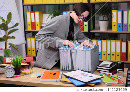 Focused office worker in gray blazer organizing documents inside storage box beside cluttered desk, surrounded by colorful binders, charts, stationery, plants, and business tools in modern workspace Focused office worker in gray blazer organizing documents inside storage box beside cluttered desk, surrounded by colorful binders, charts, stationery, plants, and business tools in modern workspace 142125669