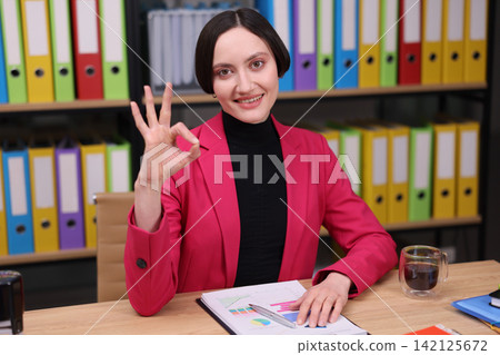 businesswoman making an ok gesture in a professional office setting with colorful binders in the background, positive and professional mood, wooden desk, paper, plastic pen, pink and dark. businesswoman making an ok gesture in a professional office setting with colorful binders in the background, positive and professional mood, wooden desk, paper, plastic pen, pink and dark. 142125672