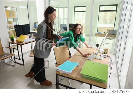 Two businesswomen collaborating in modern office, one standing with documents while coworker leans over desk and uses computer, bright glass meeting space with folders, monitors, and tidy workstations Two businesswomen collaborating in modern office, one standing with documents while coworker leans over desk and uses computer, bright glass meeting space with folders, monitors, and tidy workstations 142125674