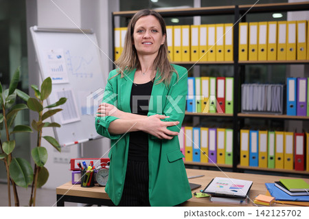 Confident businesswoman standing with folded arms in modern office beside presentation board and colorful file shelves, wearing green blazer, with focused professional expression and workspace Confident businesswoman standing with folded arms in modern office beside presentation board and colorful file shelves, wearing green blazer, with focused professional expression and workspace 142125702