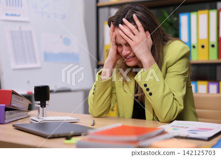 Stressed office worker holding head at desk in cluttered workspace with laptop, papers, folders, whiteboard and colorful binders while feeling overwhelmed by deadlines and heavy workload 142125703