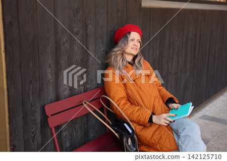Woman in red beret and orange quilted coat sitting on bench beside dark wooden wall, holding turquoise notebook and handbag, gazing away in calm reflective city street portrait Woman in red beret and orange quilted coat sitting on bench beside dark wooden wall, holding turquoise notebook and handbag, gazing away in calm reflective city street portrait 142125710