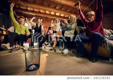 Coworkers compete to throw crumpled paper into trash bin from distance during office party. Coworkers compete to throw crumpled paper into trash bin from distance during office party. 142128191