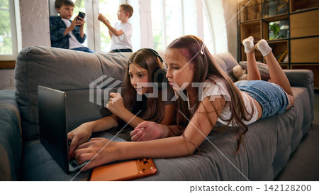 Two school-age girls browsing laptop content together while lying on sofa Two school-age girls browsing laptop content together while lying on sofa 142128200