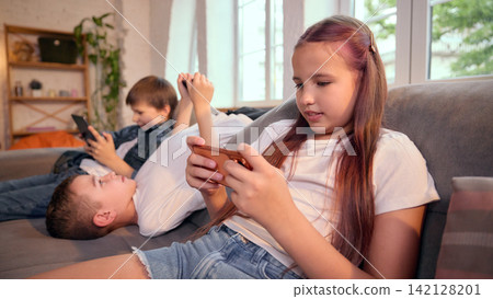 Girl using smartphone while boys play mobile games behind her at home Girl using smartphone while boys play mobile games behind her at home 142128201