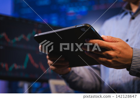 Hands holding a tablet in front of blurred stock charts and financial data screens, online trading, market analysis and business technology. 142128647