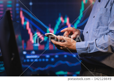 Business person using a smartphone in front of stock market charts on screens, trading, finance, and data analysis Business person using a smartphone in front of stock market charts on screens, trading, finance, and data analysis 142128649