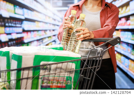 Woman shopping for cooking oil in a supermarket aisle with a grocery cart full of food and household supplies 142128704