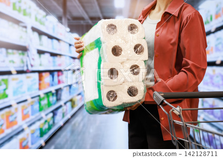 Person holding a large pack of toilet paper while shopping in a supermarket aisle next to a cart Person holding a large pack of toilet paper while shopping in a supermarket aisle next to a cart 142128711
