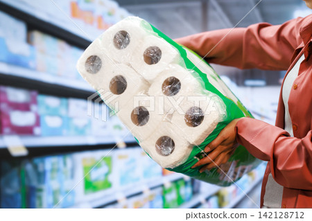 Person holding a large pack of toilet paper in a supermarket aisle, with blurred shelves in the background and a shopping scene Person holding a large pack of toilet paper in a supermarket aisle, with blurred shelves in the background and a shopping scene 142128712