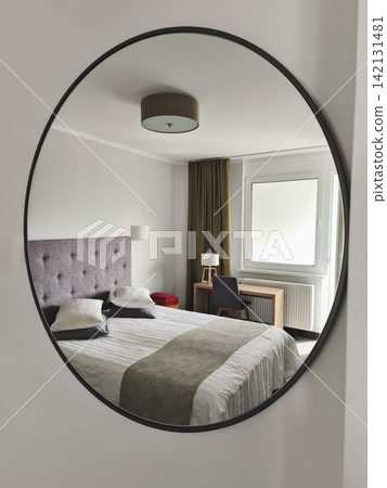 Oval wall mirror reflecting modern bedroom with double bed, upholstered headboard, window, desk and soft neutral decor Oval wall mirror reflecting modern bedroom with double bed, upholstered headboard, window, desk and soft neutral decor 142131481