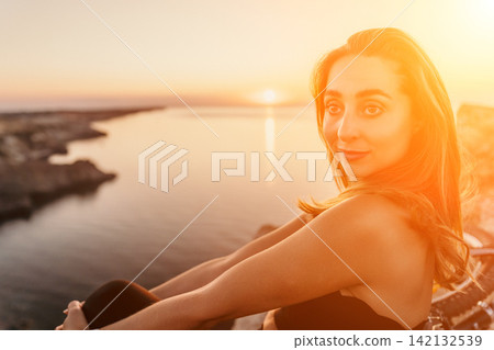 Woman sunset ocean Beautiful young woman sitting on a cliff overlooking the calm ocean during a vibrant golden hour sunset Woman sunset ocean Beautiful young woman sitting on a cliff overlooking the calm ocean during a vibrant golden hour sunset 142132539