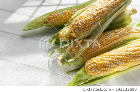 Fresh corn cobs with green husks on white wooden table Fresh corn cobs with green husks on white wooden table 142132690