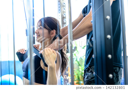 一名女子正在健身房使用下拉式器材鍛煉,並接受教練的指導。 一名女子正在健身房使用下拉式器材鍛煉,並接受教練的指導。 142132801