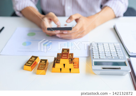 Selective focus on gold bars with background of hands using smartphone for gold trading, analyzing or managing. Concept for wealth management, asset protection, and financial planning. Selective focus on gold bars with background of hands using smartphone for gold trading, analyzing or managing. Concept for wealth management, asset protection, and financial planning. 142132872