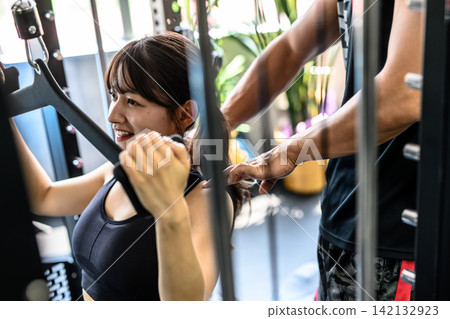 一名女子正在健身房使用下拉器材鍛煉,並接受教練的指導;白天的色彩清晰可見。 一名女子正在健身房使用下拉器材鍛煉,並接受教練的指導;白天的色彩清晰可見。 142132923